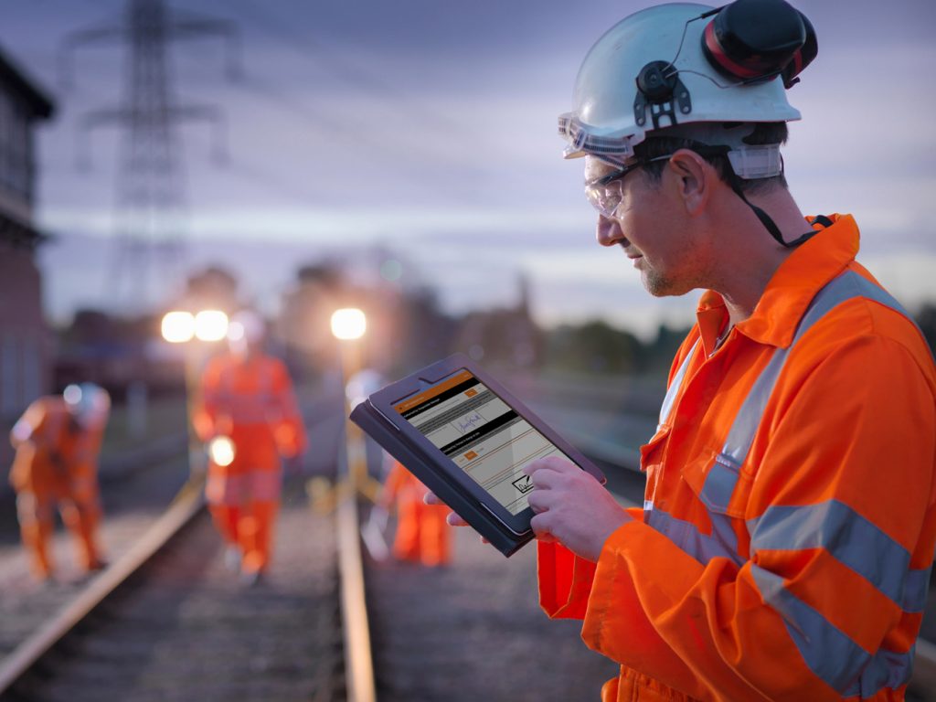 A rail worker wearing protective clothing using the ePIC app on a tablet whilst on a track side work site.